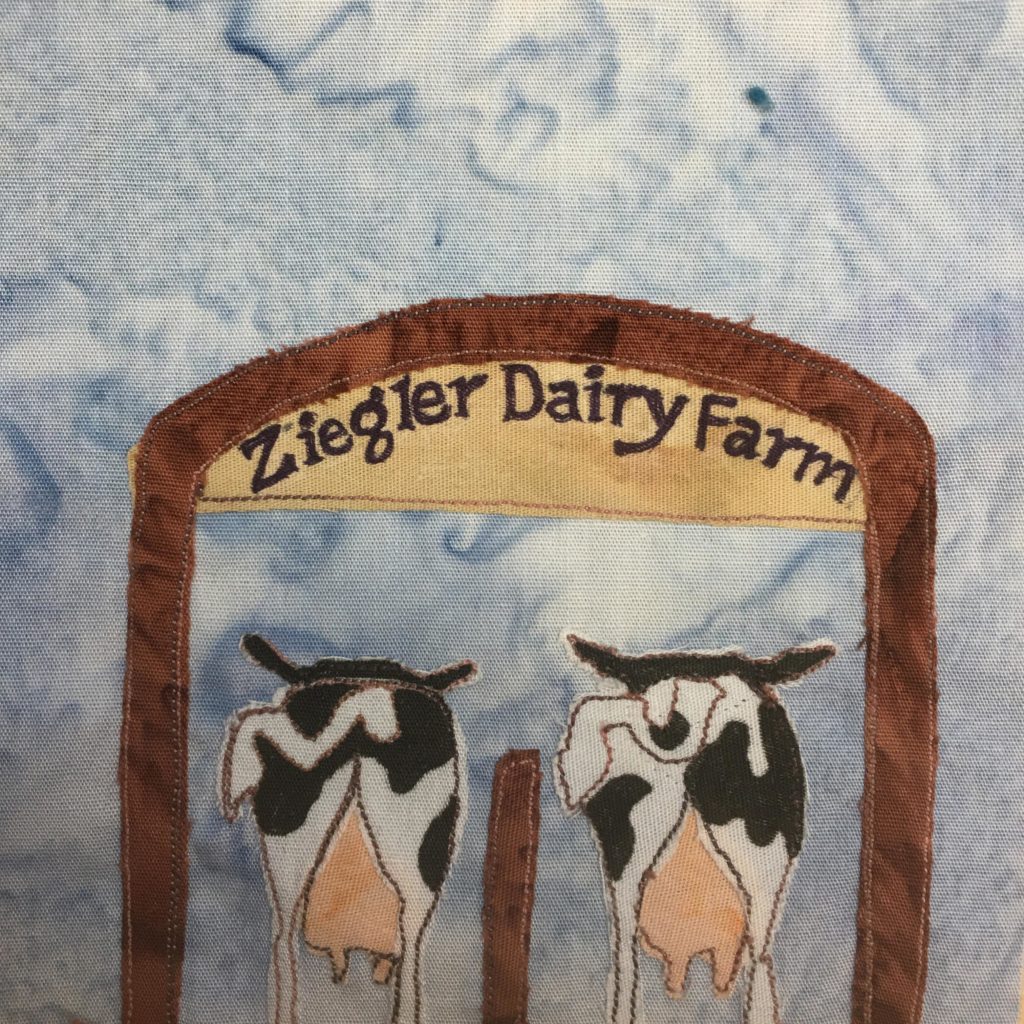 This quilt block shows two cows from behind standing next to each other with a brown divider between them. Their tails are up revealing their large udders that are pink between their back legs. The cows are pieces of white and black fabric, and look like stereotypical milking cows. There is a brown structure over them in a large arch with a tan sign beneath the arch. The sign reads,