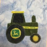 This quilt block shows a very large green tractor on top of a light blue salmon-patterned background. The tractor itself is made up of a splotchy dark green with a black fabric for the carriage window, the big tire, and the small front tire. There are yellow accents on the tractor, including its roof, a stripe on its chassis, and two yellow circles in the wells of the wheels. On the sripe in green it reads, 