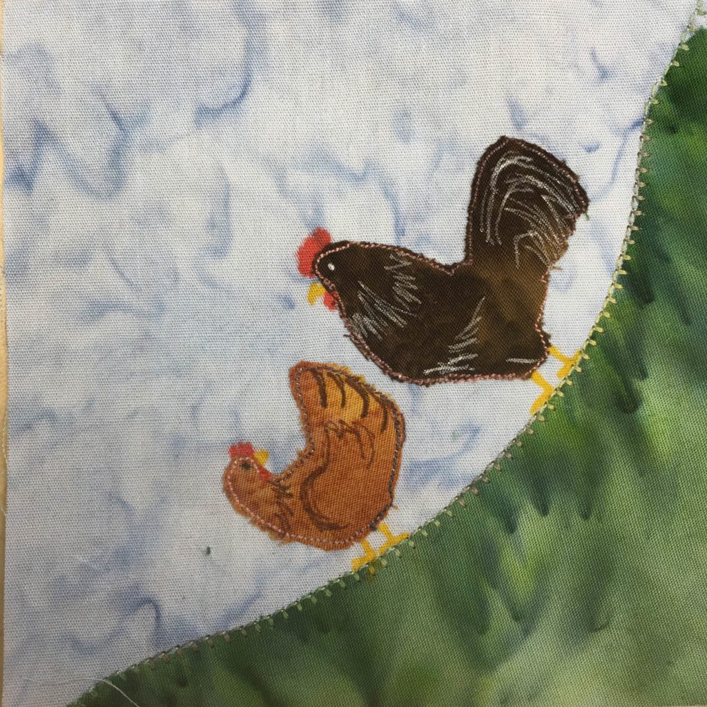 This quilt block is a corner block. There is a patch of grass in the bottom right corner, stretching from the bottom left corner to the top right corner. It has a green bold stitching across its top. There are two chickens on the grass, one a brown patch of fabric, and one a black patch of fabric. The black patch is larger than the brown patch, and is the rooster. There are pen markings on each patch to indicate feathers and a wing. The brown patch is a hen that is looking back at the rooster.
