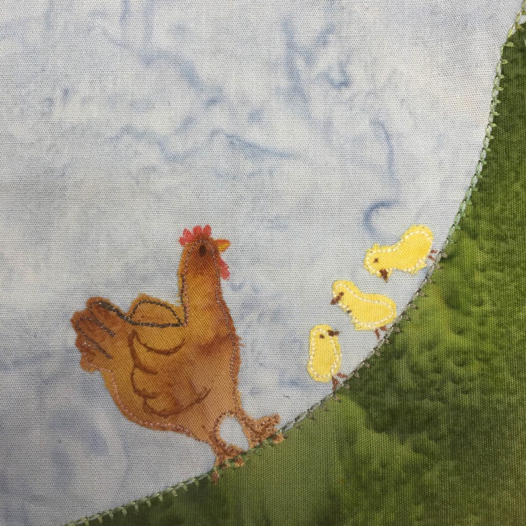 This quilt block is also a corner block. The corner is on the bottom right and is a large swatch of green fabric stretching from the bottom left corner to the upper right corner. On the curved surface of the grass is a mother hen, a cut out of brown fabric with pen marking and stitching on it to indicate the feathers and wing. Next to the mother hen are three small bright yellow patches of fabric shaped like baby chicks. Each baby chick has a little beak and eye in pen, and two tiny feet.