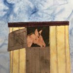 This quilt block shows a horse in a stable. The stable is a yellow building with vertical slats delineated by red stitching. In the middle of the building is a brown gate split into an upper window and a lower door. The upper window is open, and out from the black fabric inside is an orange fabric in the shape of a horse. The horse has extra details drawn in with pen, like an eye and mane. The roof of the stable is a dark red and flat.
