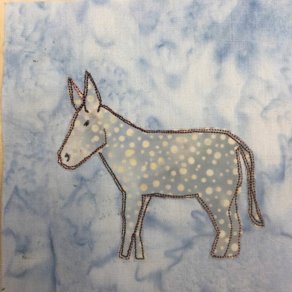 This quilt block, much like the last one, shows a donkey or mule against a light blue background. The mule is made up of a polkadotted duotone gray-yellow fabric. It is outlined by a black tight stitching. You can see the mule's large ears and much broader and longer head. The mule also stands shorter than a regular horse with stocky back legs. It also has a long thing tail with a little bush at the end.