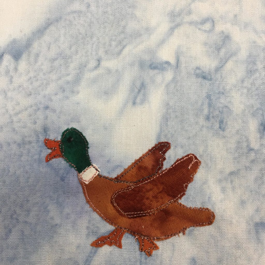 This quilt block shows a duck from the side facing left. The duck has a green head, a white collar, and a brown body. The duck bill is a small orange piece of fabric, the head a dark green piece of fabric, the body a small white piece, and the body is made up of three brown pieces (two for the wings). The feet are the same fabric as the bill. The wings are in mid-flap and are coming out of each side of the duck. Every piece of fabric is outlined by a black and white salt-and-pepper-like stitch. This is against a light blue salmon-patterned background.