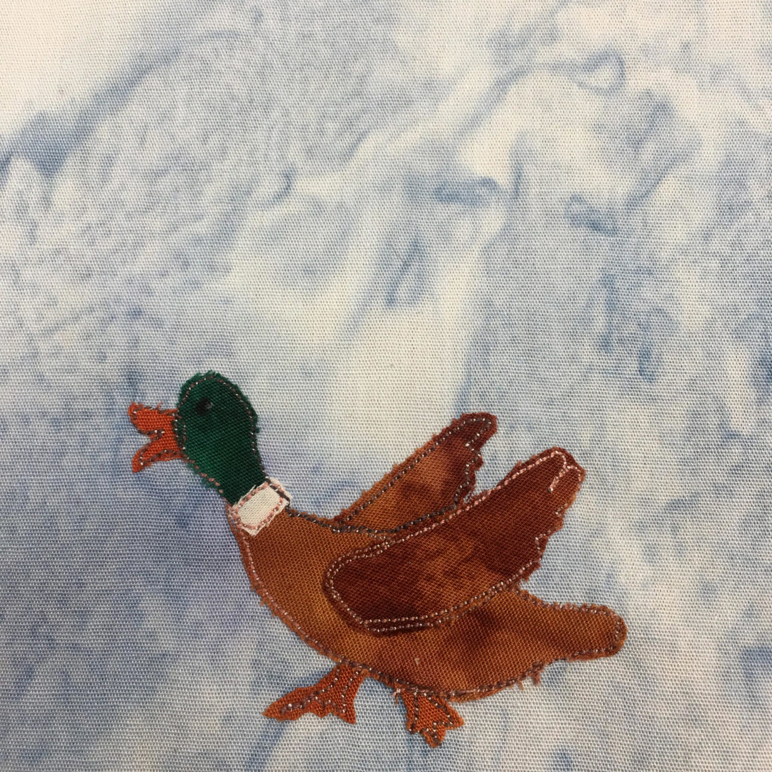 This quilt block shows a duck from the side facing left. The duck has a green head, a white collar, and a brown body. The duck bill is a small orange piece of fabric, the head a dark green piece of fabric, the body a small white piece, and the body is made up of three brown pieces (two for the wings). The feet are the same fabric as the bill. The wings are in mid-flap and are coming out of each side of the duck. Every piece of fabric is outlined by a black and white salt-and-pepper-like stitch. This is against a light blue salmon-patterned background.