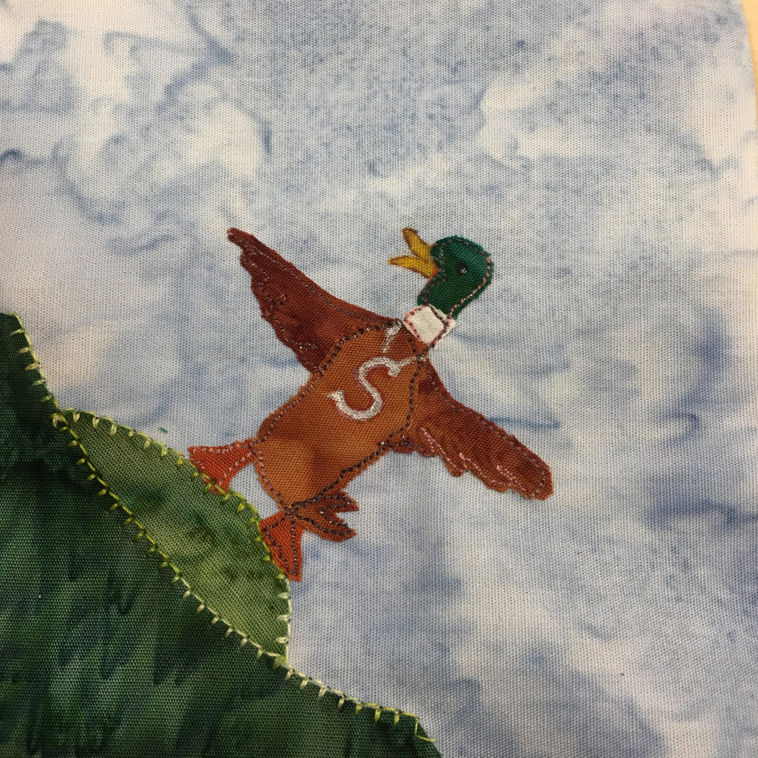 This block shows a patch of corner grass with the strong stitching in light green on its border. This patch of grass is two dark green fabrics, one a mound on top of the other in the bottom left corner. Standing on the mound is a duck, just about standing up flapping and stretching its wings. On the breast of the duck is a large white S written in with white pen. The duck is a several colors. The bill is a yellow fabric, the head a deep green fabric, the neck a white fabric, and the rest (beside the feet) is a brown fabric. The feet are orange fabric. These are all stitched together with a tight salt-and-pepper stitching to look very much like a duck standing up.