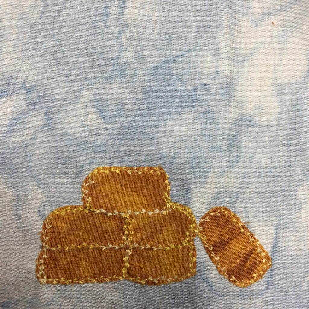 This quilt block shows a stack of straw bales. The bales themselves are a very yellow orange fabric. Each bale is outlined by a very frilly leaf like stitch of very gold thread. There are four bales in a block, with one bale on top. There is another bale to the right leaning on the main pile of bales.
