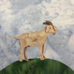 This quilt block shows a majestic wether goat standing on a mound of grass. The mound is a dark green fabric with no stitching. On top of the mound is a tan fabric in the shape of a goat. The back legs are shaded in with pen. The goat's head is outlined with stitching and with brown pen. The horns are a black fabric with some white stitching on them.
