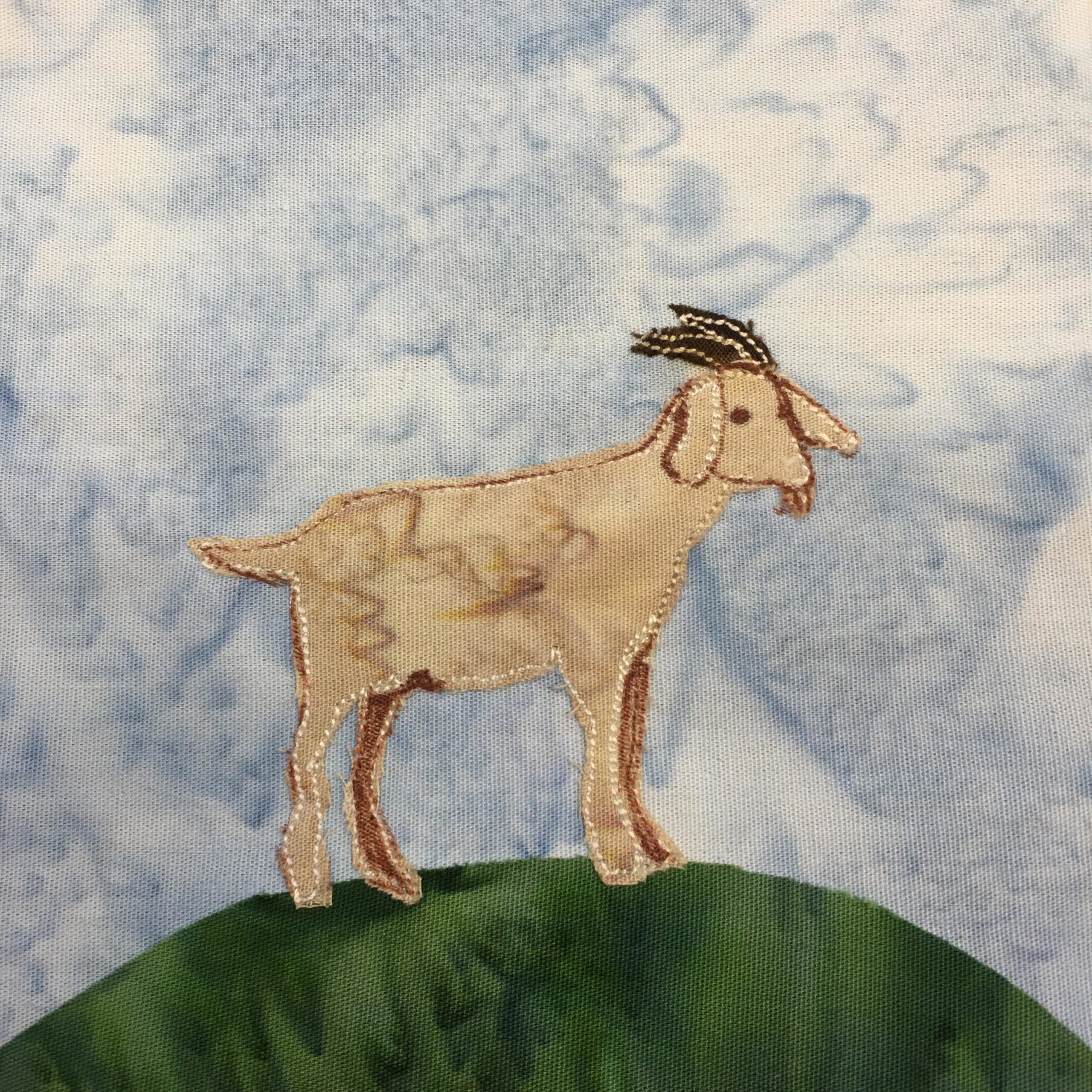 This quilt block shows a majestic wether goat standing on a mound of grass. The mound is a dark green fabric with no stitching. On top of the mound is a tan fabric in the shape of a goat. The back legs are shaded in with pen. The goat's head is outlined with stitching and with brown pen. The horns are a black fabric with some white stitching on them.