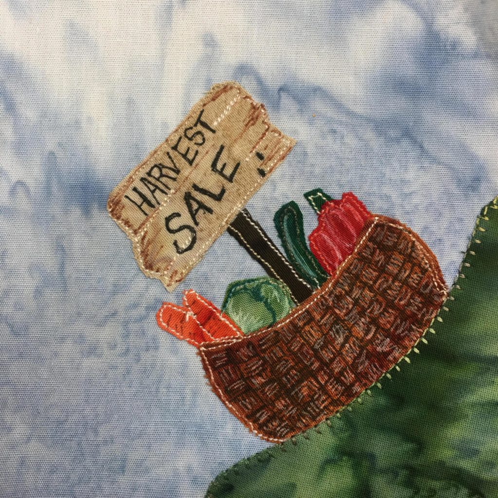 This quilt block is a corner block. it has a dark green patch of grass in the bottom left corner. On that patch of grass is a basket. The basket is a brown fabric with a lot of pen marks on it in a hatched design to make it look like a weaved basket. Inside the basket are several vegetables. There are carrots, cucumbers, lettuce, and a red pepper each with corresponding colored fabric. Coming out from the center of the basket is a stick that's holding up a detailed sign that has written in black, "Harvest Sale."