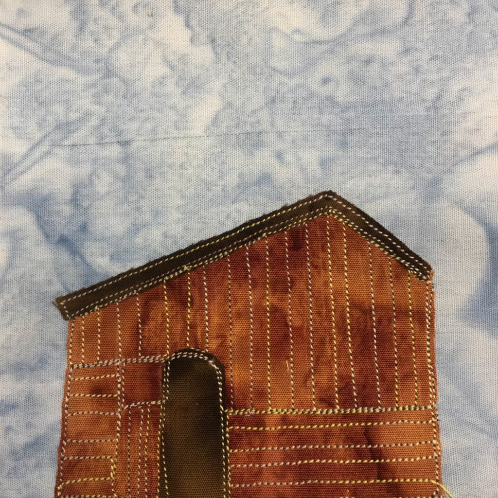 This quilt block shows a brown building made out of wooden slats. The slats are shown with yellow stitching. There is a door on the left side that is a very dark brown color. The roof is the same color and is slanted, with the top point of the roof to the right (the left side is longer than the right side). This is against a salmon-patterned blue background.