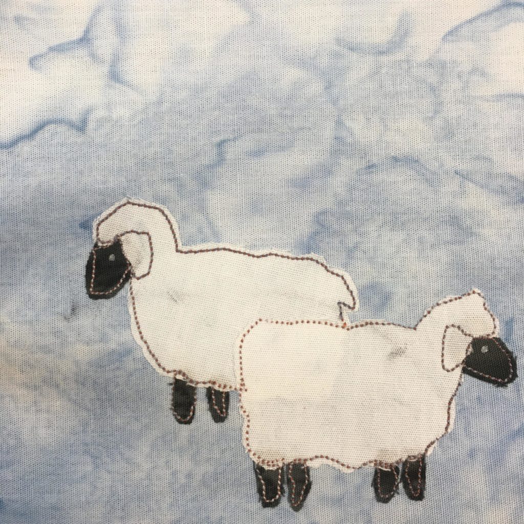 This quilt block shows two black skinned and white wool sheep against a salmon-patternd light blue background. Each sheep is a large patch of white fabric that is shaped somewhat in a cloudy, or woolly, way. Each sheep's ear is delineated by stitching inside the white. Poking out of the ends of the tops are black pieces of fabric to indicate their faces. At the bottom of each woolly piece of fabric are short black pieces for their legs. Each piece of fabric is outlined with a light brown stitching.