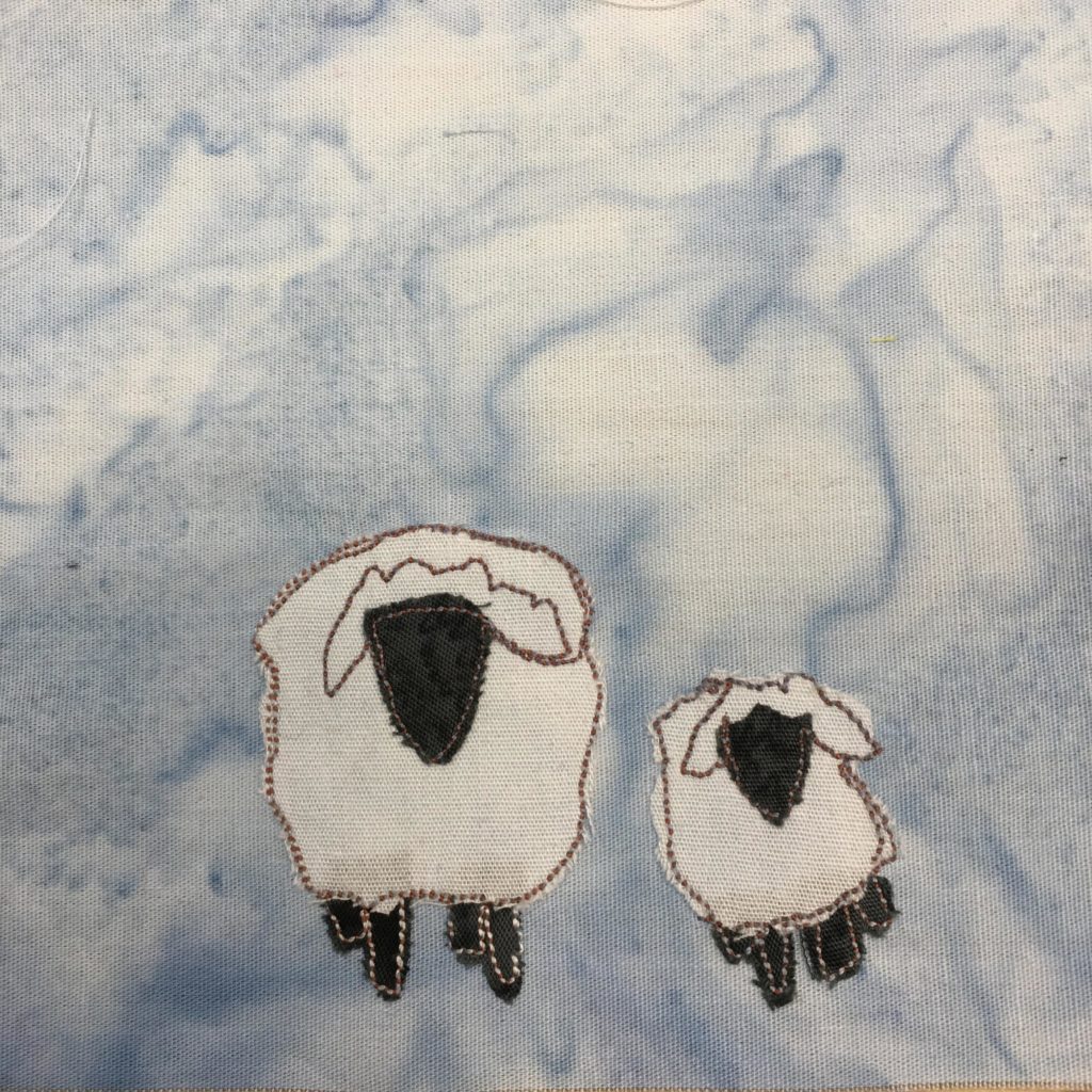 This quilt block shows two sheep from the front. One sheep is much larger than the other, and is the mama sheep to the lamb. Each sheep is a piece of white fabric with a black triangular face near the upper middle. The bodies and ears are outlined with a brown threaded stitching. Each sheep also has four black pieces of fabric indicating their short legs.