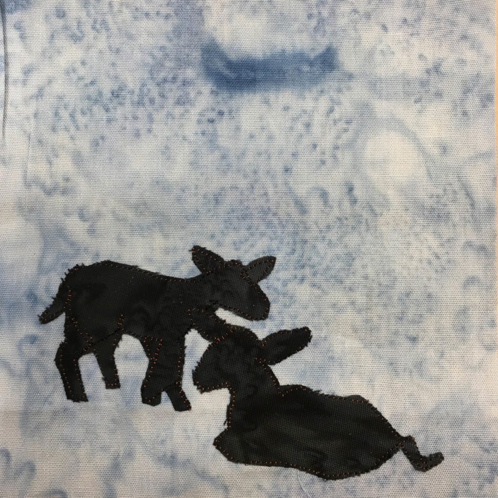 This quilt block shows two young Suffolk lambs against a light salmon-patterned blue background. The lambs themselves are made up of shaped black pieces of fabric, like silhouettes. One is shaped in such a way that it is standing and taking a step, while the other is laying down with its legs folded underneath itself. The black pieces of fabric (and the ears and other details) are outlined with a very small orange fabric stitching.
