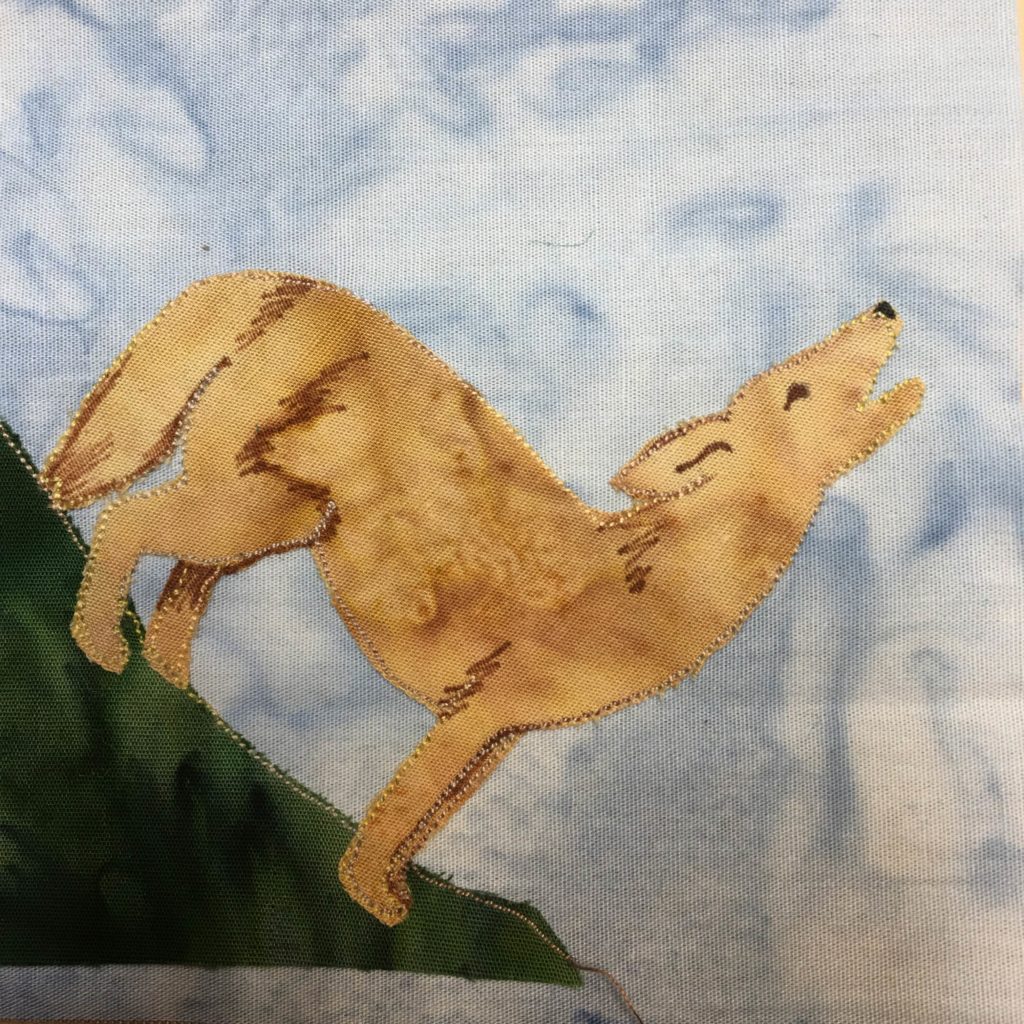 This quilt block is a corner block. This means that there is a patch of grass in the lower left corner indicating the ground. The other corners are open. Against a blue salmon-patterned background is a dark green piece of fabric, and a tan salmon-patterned piece of fabric. The tan fabric is in the shape of a coyote. The coyote is detailed further with pen markings indicating the shape of the legs and furry tail. The coyote is howling, with its head going straight up and its mouth open.