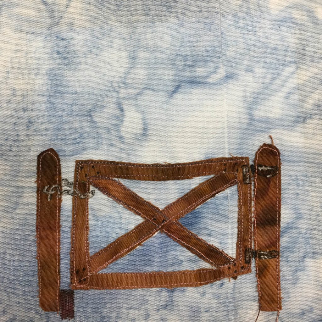 This quilt block shows a large brown gate against a blue salmon-patternd background. The gate has two posts on on either side of it. The gate itself is a rectangle frame with an x through it with two extra bars. Below the left side of the gate is a block to prop the end of the gate on to keep it form sagging. There is a chain and hinges drawn on the brown fabric in pen.