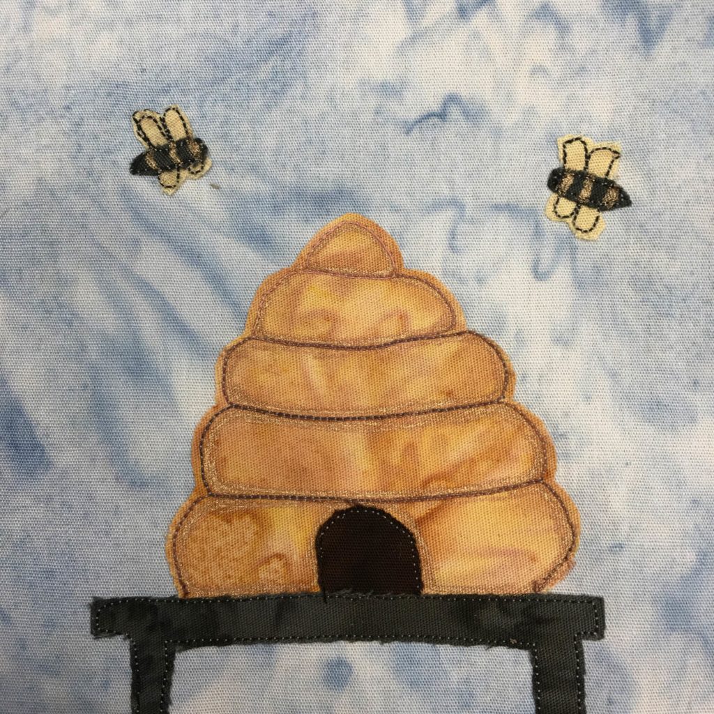 This quilt block shows a very stereotypical beehive with two large bees flying around it. The beehive looks like the hairdo, being a stack of smaller rolls laid on top of each other. The hive is a honey color, and the bees are yellow with their stripes penned in (and pretty big wings)! The hive is standing on a dark gray table.
