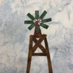 This quilt block shows a windmill structure against a light blue salmon-patterned background. We're seeing the windmill top. There's a brown structure holding up the top that is a stack of brown wood leaning on each other with an X structure for support. The top of the windmill has a deep red triangle pointing upwards with a big circular fanout of bluish-green slats attached to the tip of it like a wheel. Each piece of fabric, including the slates is outlined with a light brown stitching.