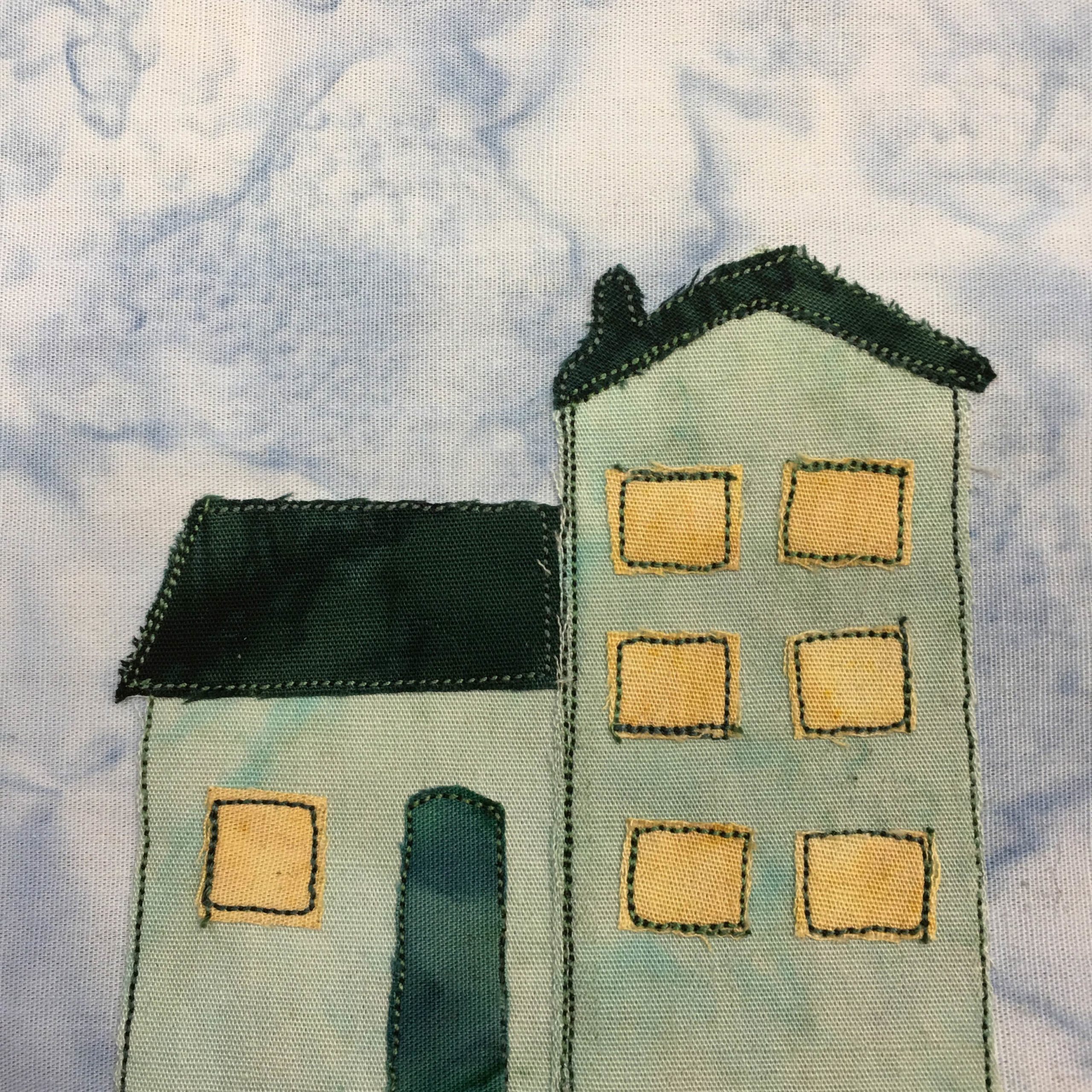 This quilt block shows a light green house on a light blue salmon-patterned background. The house is in two sections, one next to the other. The left section has a dark teal door with one yellow square window and a slow very dark green roof. The right section is three stories tall (at least it has three rows of square windows going up.). It's roof is facing away from us and is in the shape of a triangle. There's a little chimney in this roof. The windows are a light yellow with green stitching.