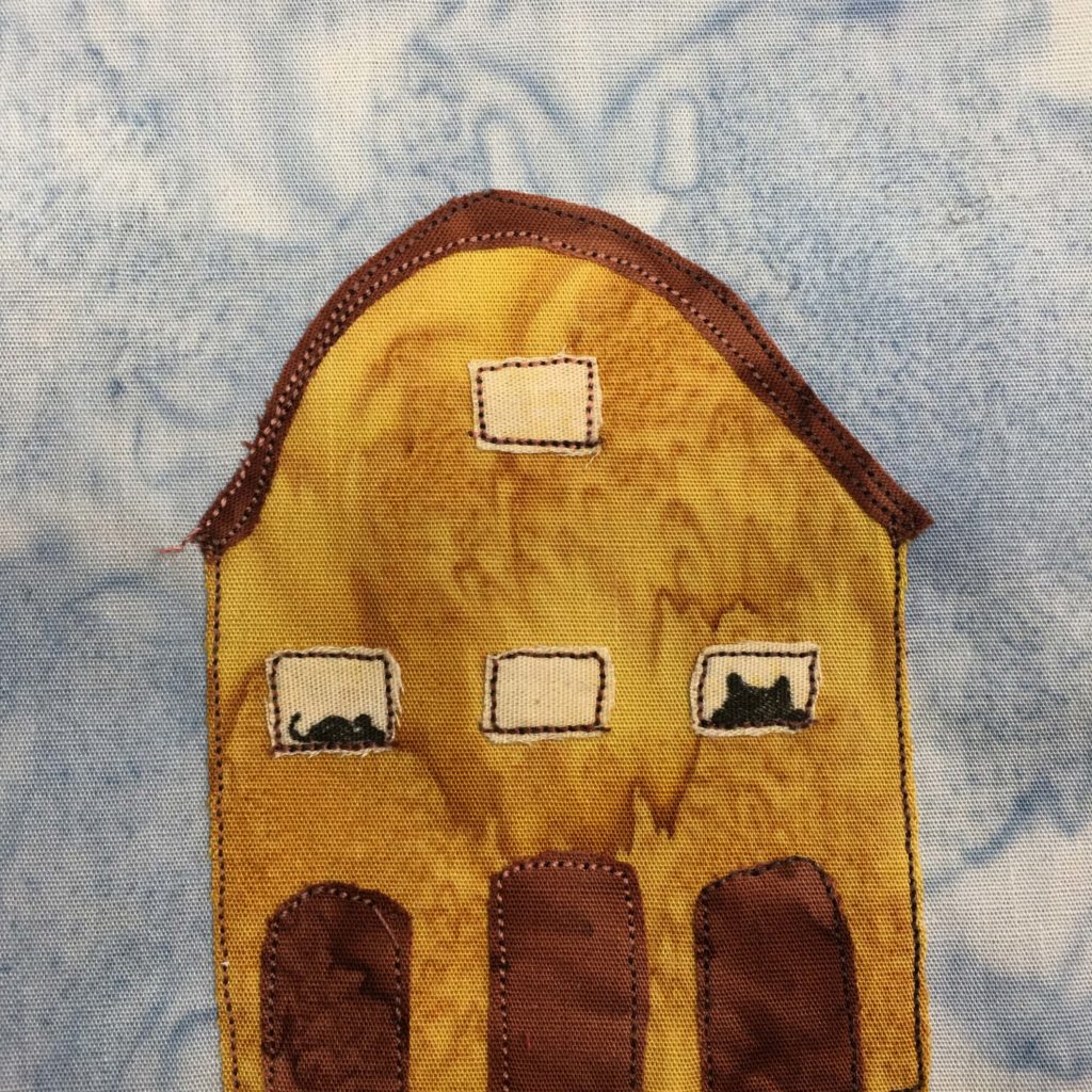 This quilt block shows a tall orange building with a roundish roof. It looks like a big barn on a farm. There are three entry doors on the bottom, all made out of deep red pieces of fabric. There are three windows, each above one entrance. In the left window you can see the black silhouette of a mouse, and in the far right window you can see the silhouette of a cat's head. The roof is the same deep red as the doors.