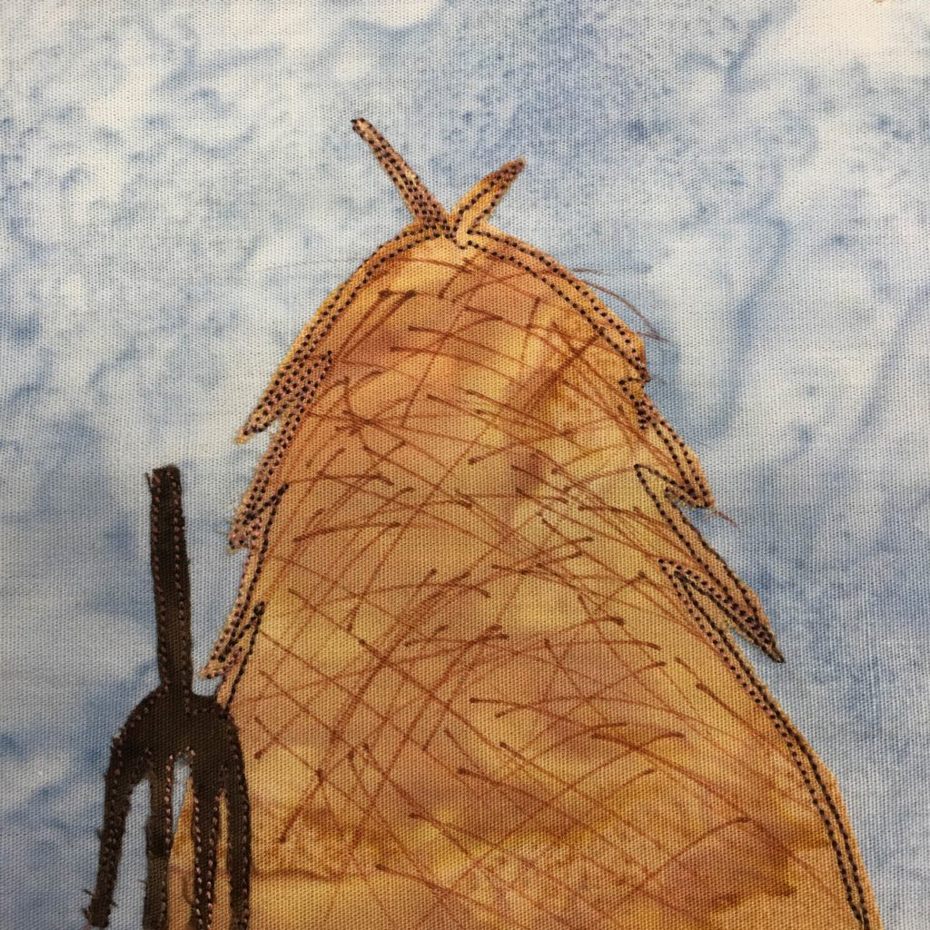 This quilt block shows a huge hay pile. The hay pile is a large piece of yellow-gold fabric with darker orange pen markings on it. It's outlined with a deep brown stitching, the same color as the piece of fabric making up the pitch for to the front and left.