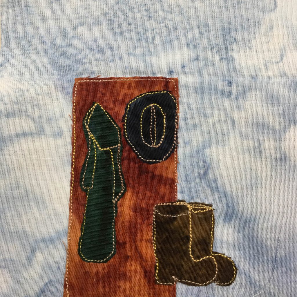 This quilt block shows a door, or a panel of wall that would be next to a door, with some pegs for hanging things. The pegs can't be seen because a large brimmed hat made out of black fabric and a cloak made out of a dark green fabric are hanging from the wall. Next to the wall on the bottom right are two gray boots. Everything is outlined in a bright yellow stitching.