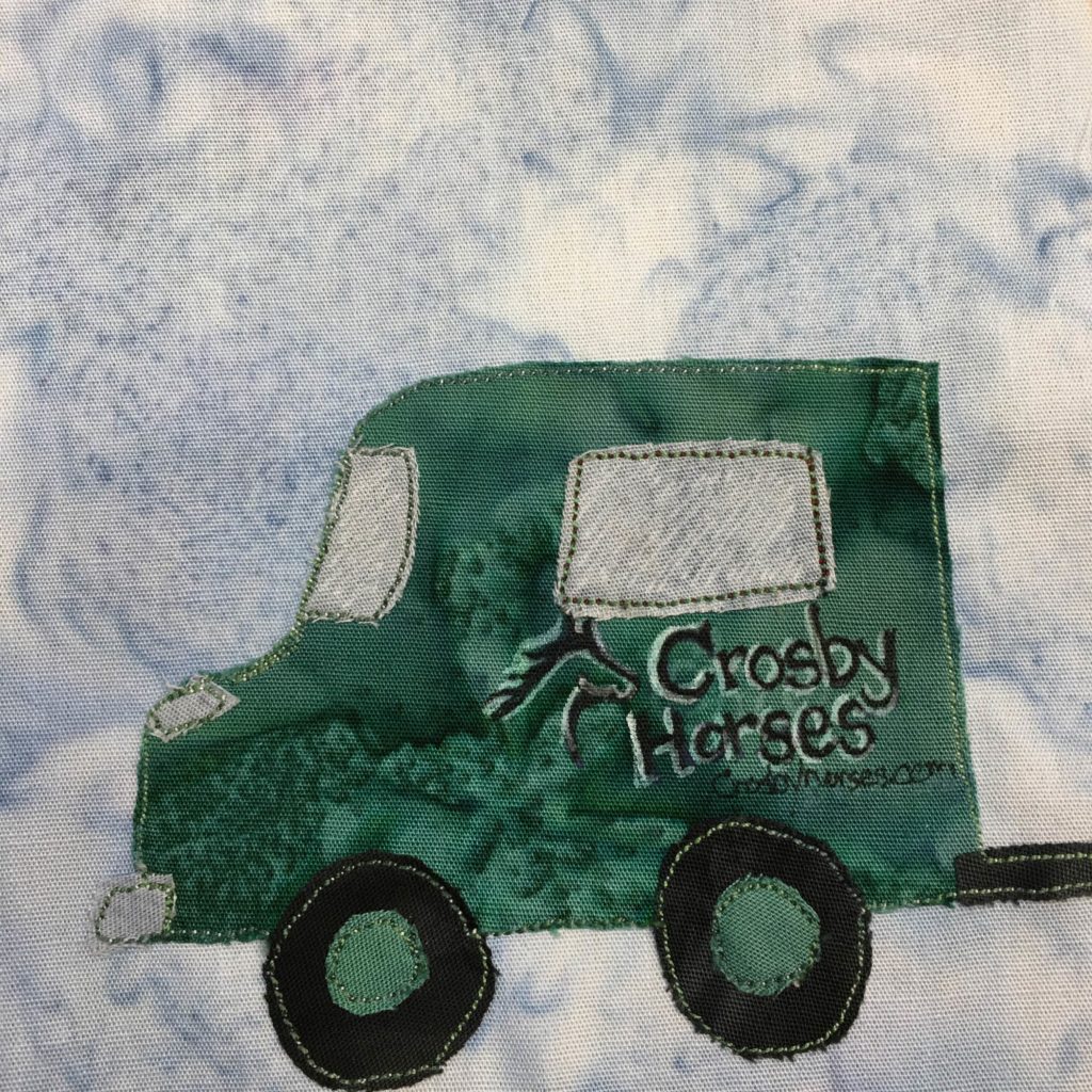 This quilt block shows a very green SUV looking car from the side. It's bumper in the back looks like it's towing something. The wheel axles are green, while the wheels are black pieces of fabric. The side of the truck has a pen drawing with white accents that say, "Crosby Horses." The windows are gray and have white pen lines on them to show the reflection.