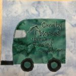 This quilt block shows the trailer attached to yesterday's SUV. The trailer is the same tint of green that the SUV was, which is a darker green. On the side is a big logo written in black and white pen (white to highlight the outlines) that shows a horse head and neck and the text, "Crosby Horses • Breeding • Training www.crosbyhorses.com" There are two sets of wheels on the trailer, despite it's short length, which are black with green axles. There isalso a window on the upper front of the trailer which is a white fabricwith white pen lines showing the reflection.