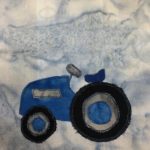 This quilt block shows a blue tractor from the side. It's very blue. The blue fabric is accented by a silver fabric. The silver fabric makes up the inside of the wheels, the steering wheel, and a small stripe on the side of the chassis. The wheels are a black fabric while the hubcaps are blue. The tractor is a smaller tractor with a little open seat in front of the steering wheel, and one big tired behind a small one.