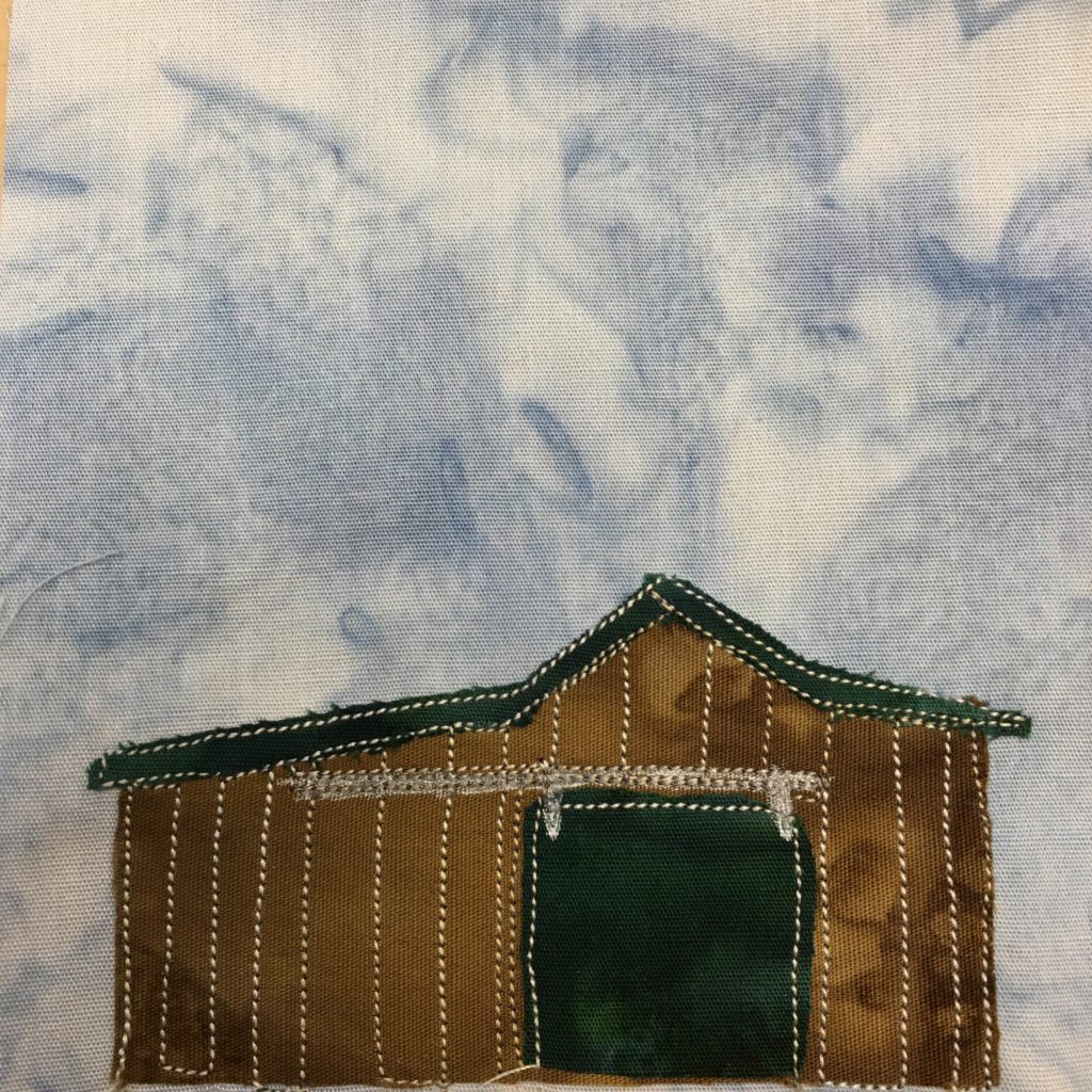 This quilt block shows a structure, a building, with one side larger than the other. It's a squat brown building with slats of wood making up the walls. These are indicated by white stitching outlining each slat. There is a giant sliding wooden door with a rail jutting out to the larger side for it. The sliding door is a dark green, along with the roof of the shed. This is all against a light blue salmon-patternd background as usual.