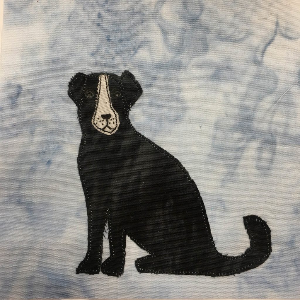 This quilt block shows a black and white dog against a light blue salmon-patterned background. The dog is made up of a cut out piece of black fabric, with a white stripe of fabric descending from her head and making up her muzzle. The dog is sitting and looking towards us. On each side of the head, with the stitching you can see two ears, and with a little pen you can see the two black eyes.