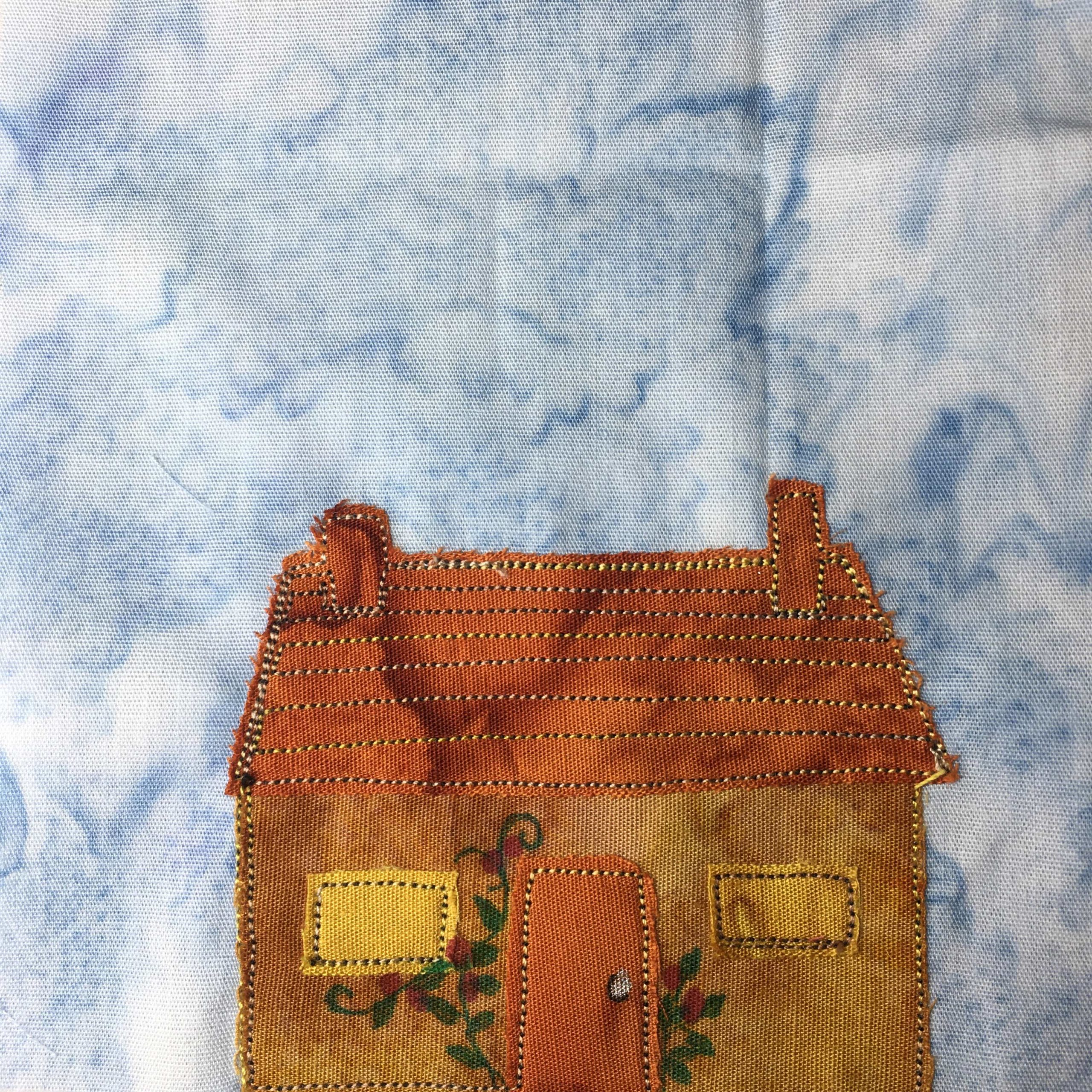 This quilt block shows a lovely orange cottage in the bottom half. The cottage has an orange roof, with two chimneys. The front is a darker orange tan with two square golden windows. The door is orange like the roof and has a white doorknob. Penned in on the front of the house is a green vine with red flowers on it, traveling up the wall. The roof is lined with stitching and the house and windows are outlined with stitching.