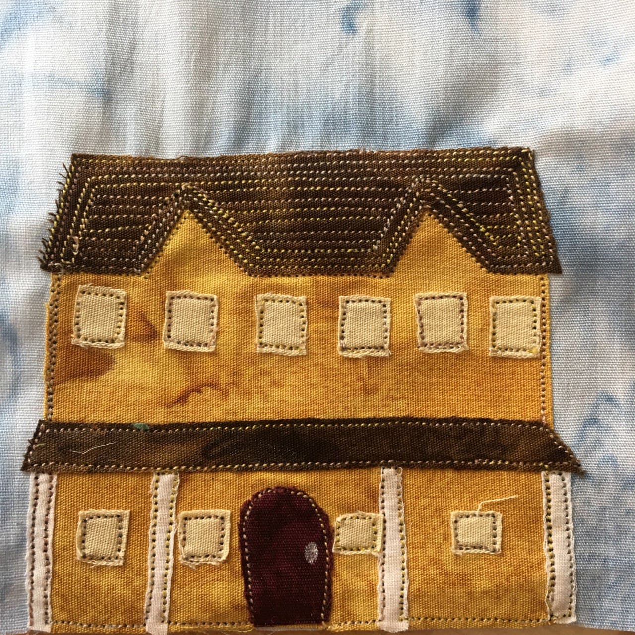 This quilt block shows a very large house against a blue background. The house is two-storied, with two peaks in the slanted roof that's coming down towards the viewer. The upper floor has six square yellow windows, each outlined by a black stitching. The lower floor has an awning coming out that's supported by white pillars. The awning and the roof are the same color dark brown. Behind the white pillars, also outlined in black, are four square yellow windows. These windows are two on each side of a round dark brown door with white stitching. It's indeed a large hosue.
