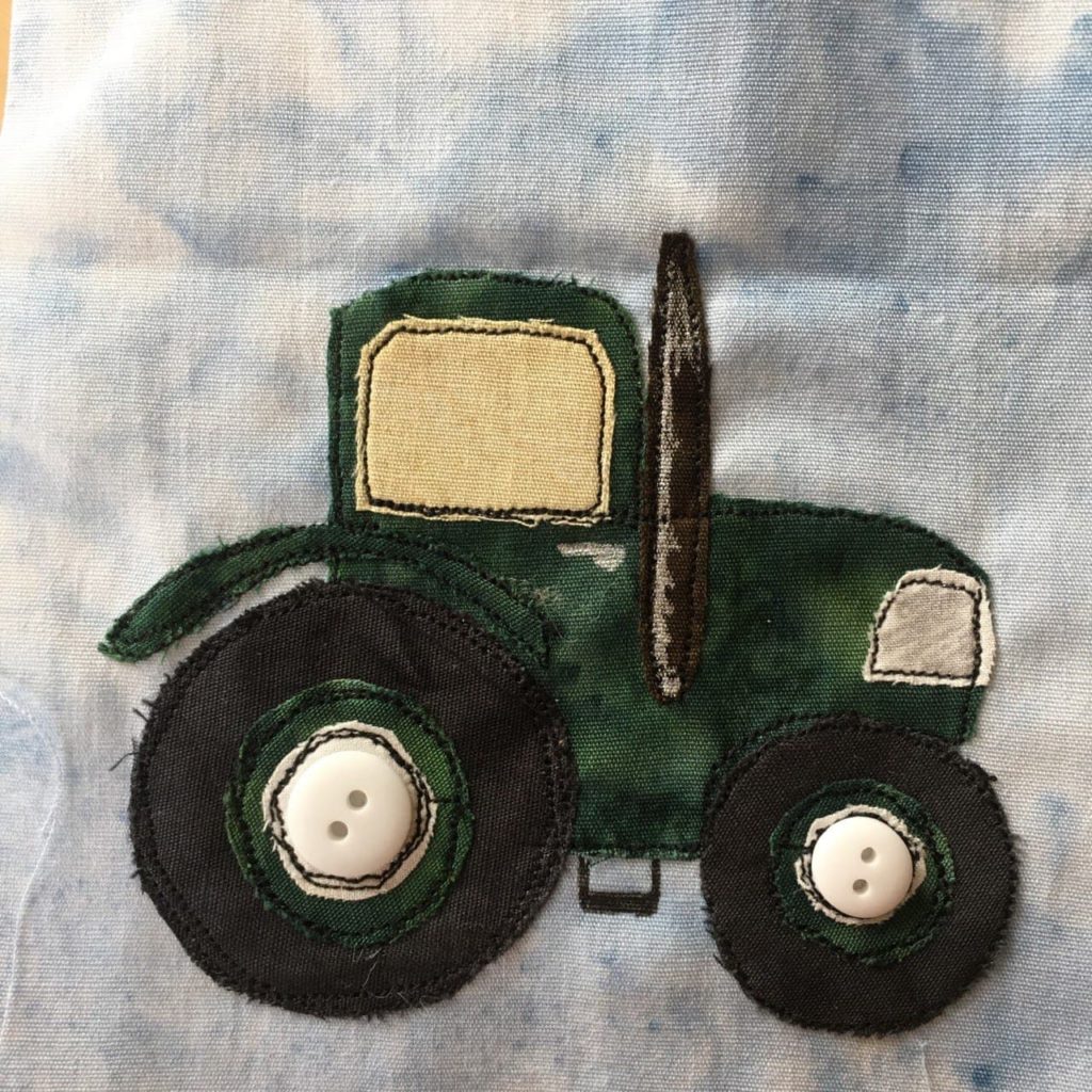 This quilt block shows a fancy dark green tractor. It has a large back wheel and a smaller front wheel. There's a black smoke stack the same color as the wheels on the side and extending above the cab. There is a large white headlight on the front of the tractor (we're viewing it from the side) and a large cab with a light yellow tinted window. The smoke stack has white pen on it to show it's sheen. There are two white buttons in the axles of the wheels, one larger than the other. To finish it off in pen there's a small step below the chassis to help you into the tractor's cabin.