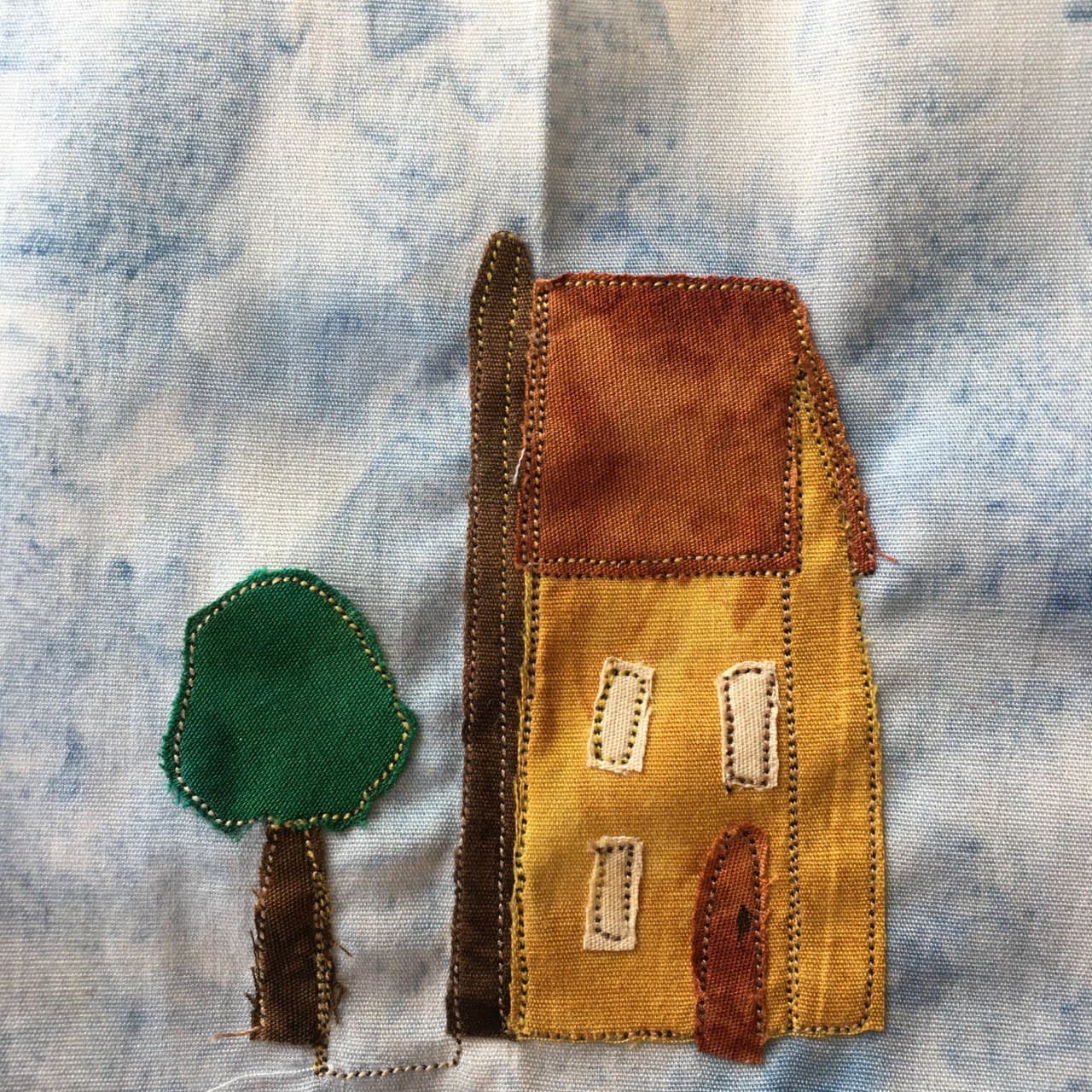 This quilt block shows a funny shaped house next to a tree. The house has a bit of an odd curve shape on the side that helps make it look like it's turning towards the viewer. The roof of the house is sloped and is a light brown, while the body of the house is a light rusted tan. There are two floors with windows, each window an offwhite rectangle of fabric. There is a thin door that is a darker brown. On the left side of the house is a dark brown tall chimney that goes from the bottom ad above the roof. The tree has a rich green foliage, being a piece of green fabric. There is stitching outlining each window and the house, as well as the tree, a black and white pepper colored tight stitching.
