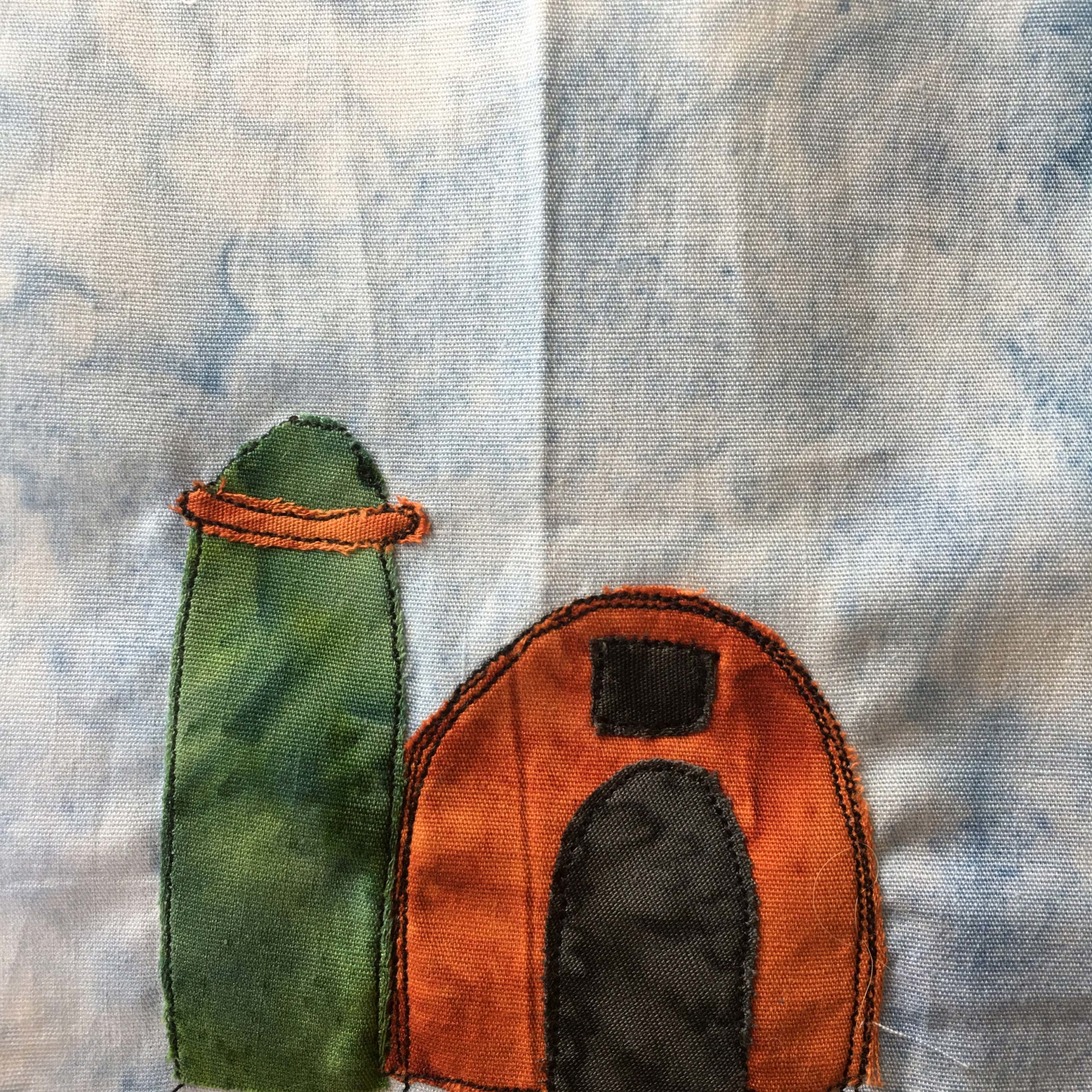 This quilt block shows a tall green grain silo next to a round barn like building that's orange. The entrance to the orange building is a large black archway, and there's a black window above it. There is a dark orange, same colors s the building, ring around the top of the grain silo on the left. Pieces are stitched with a black thread.