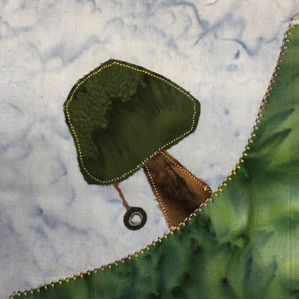 This quilt block is a corner block. This means that a piece of green fabric representing grass stretches from the bottom left corner to the top right corner. The grass is topped with a thick yellow stitching. On top of the grass is a tree with a cone shaped trunk made of dark brown fabric, and a large foliage on top made of a dark green fabric. These are outlined by a thinner yellow stitching. There is a rope and tire hanging from the tree drawn in with pen.