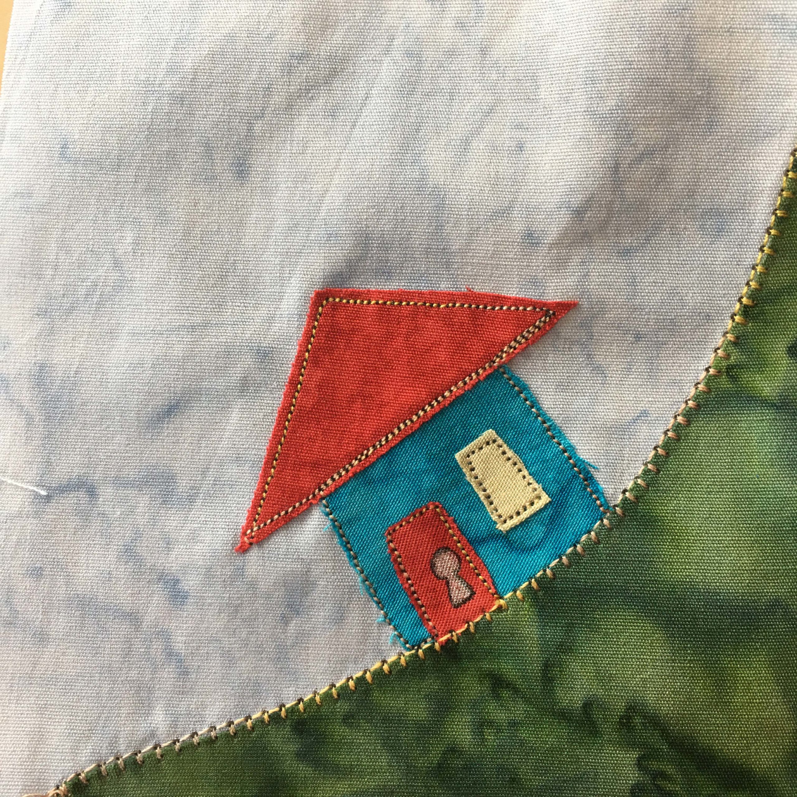 This quilt block is another corner block. This means there is a piece of green fabric signifying grass that stretches from the lower left corner to the upper right corner. It has a strong yellow stitching on it's edge. Sitting on the grass is a brightly colored house. It has a red triangular roof, a blue body, and a yellow window. There's also a red door with a large keyhole drawn in using pen.